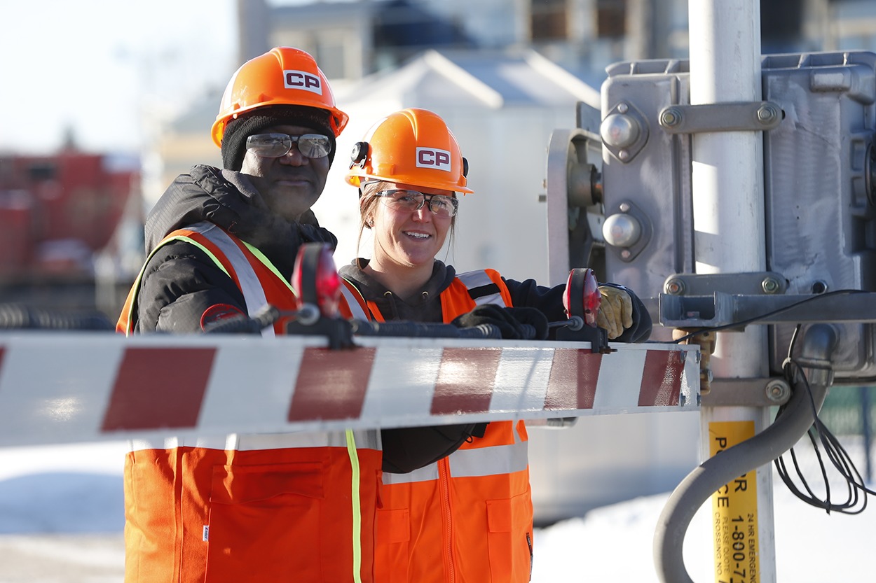 Two employees wearing safety equipment standing at a rail crossing guard.
